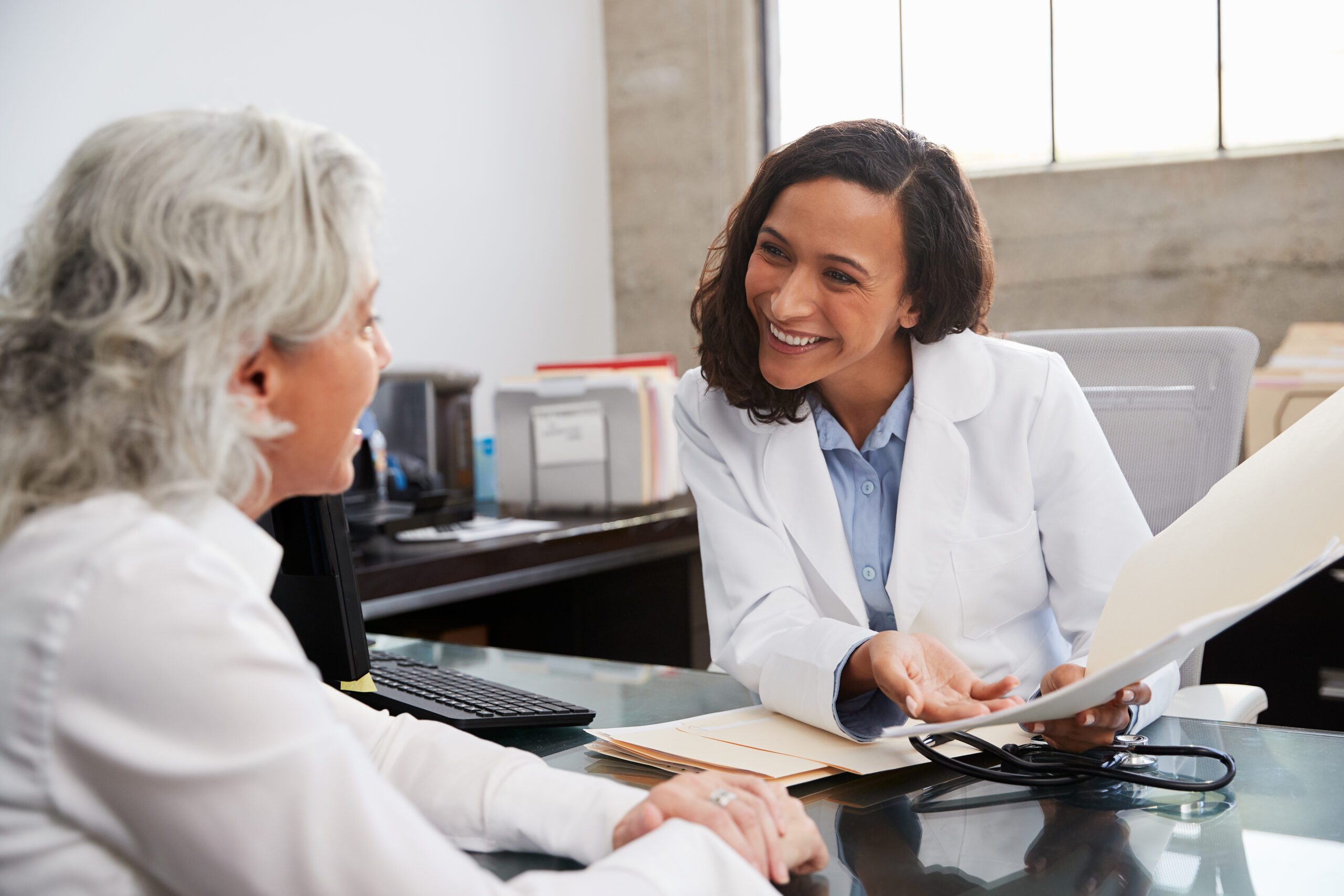 A photo of a doctor wearing a white coat, smiling at a female patient sat across the desk from her