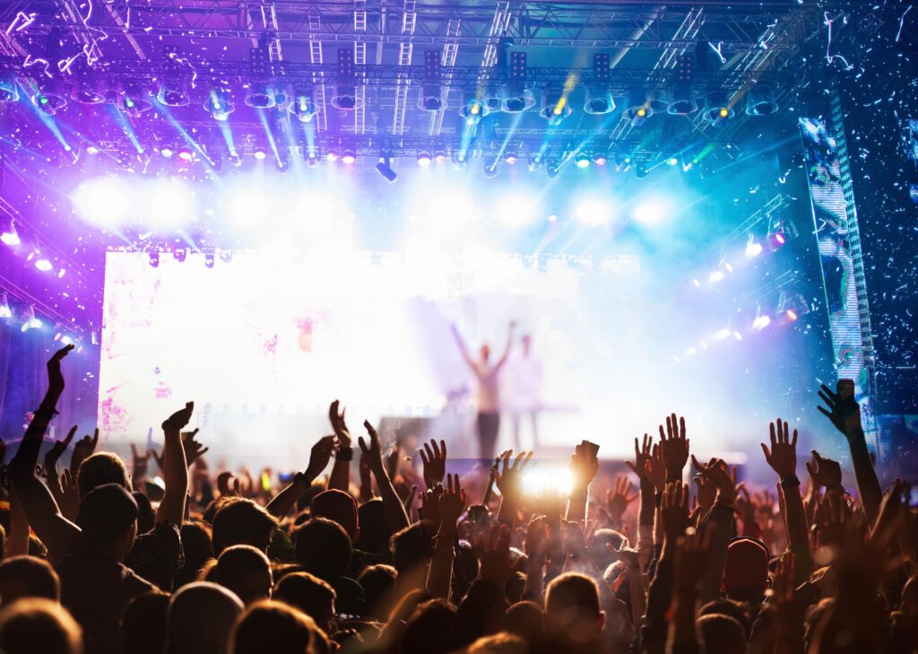 A photo of a live music event taken from behind the crowd, where people are putting their hands in the air and there is a full view of the stage lit up with bright lights, where a singer stands with their arms raised above their heads