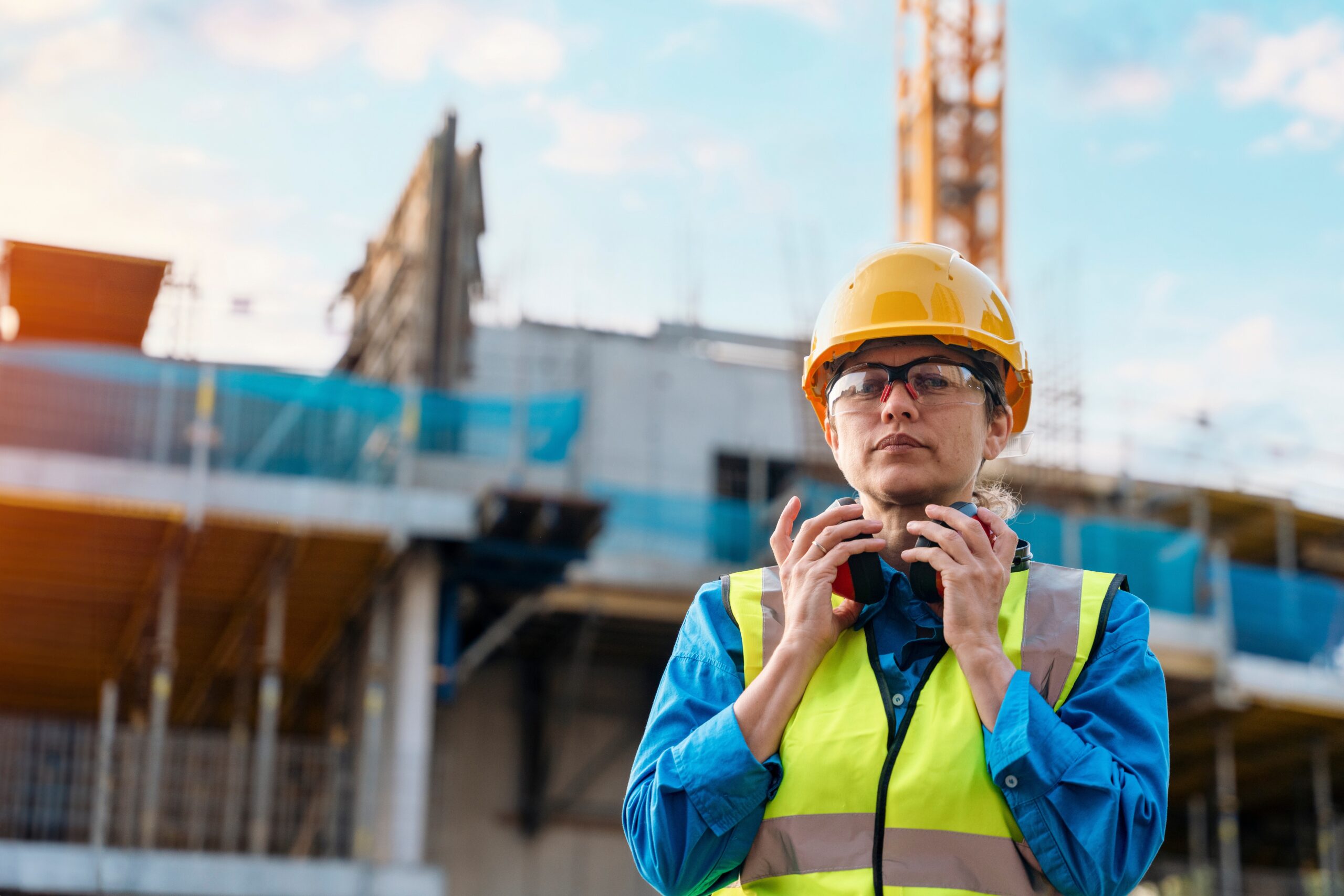 A female construction worker stands wearing a high vis vest holding ear defenders that are resting around her neck.