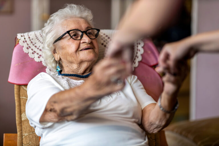 An elderly woman is sat in her armchair with her hands outstretched and is holding hands with a carer who is stood in front of her but not seen by the camera