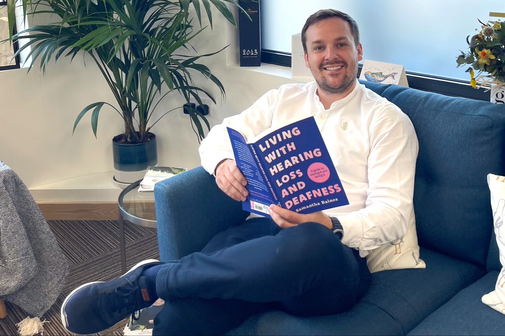A man is sitting in a chair, holding a book in his hands about living with hearing loss and looking up and smiling at the camera.