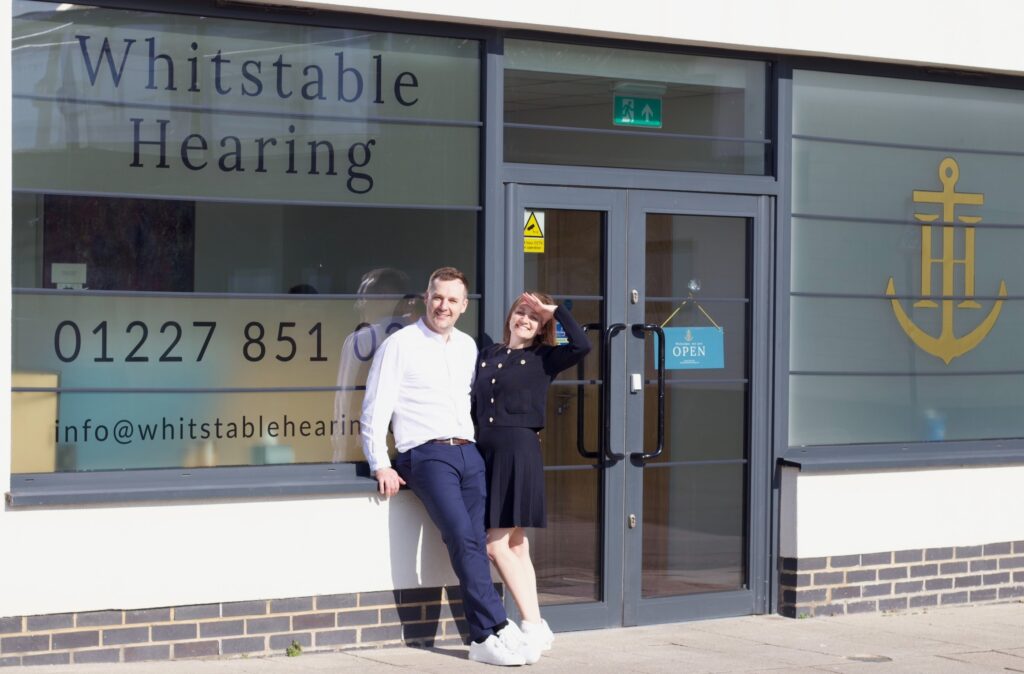 Adam and Amy Chell stand outside the front of Whitstable Hearing, their audiology clinic, leaning against the window with their arms around each other.