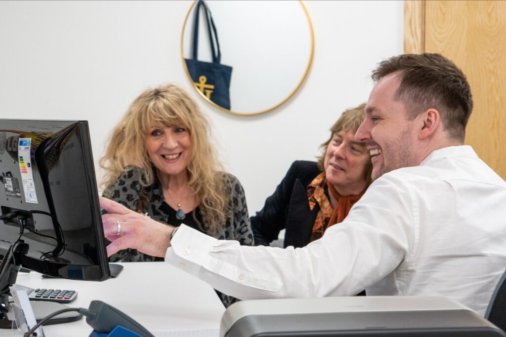 Three people sit around a computer screen. A man is gesturing to something on the screen and they are all smiling.