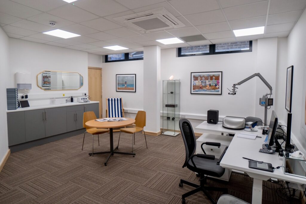 A photo of an audiologist's clinic room, showing a desk with a computer on it and a chair on one side of the room, a small table ans two chairs in the centre and some cupboards and equipment against the other side of the room. There is a deckchair in one corner and two photos on the back wall.