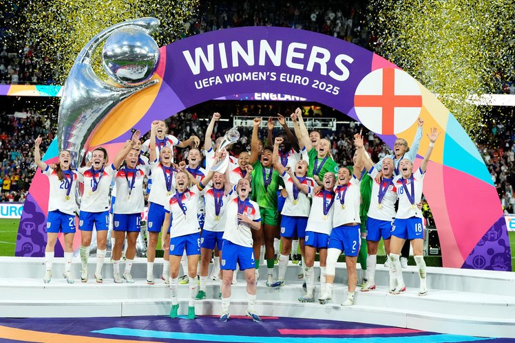 The whole England women's football team stand together on a stage, jumping, cheering and celebrating their Euro 2025 tournament win