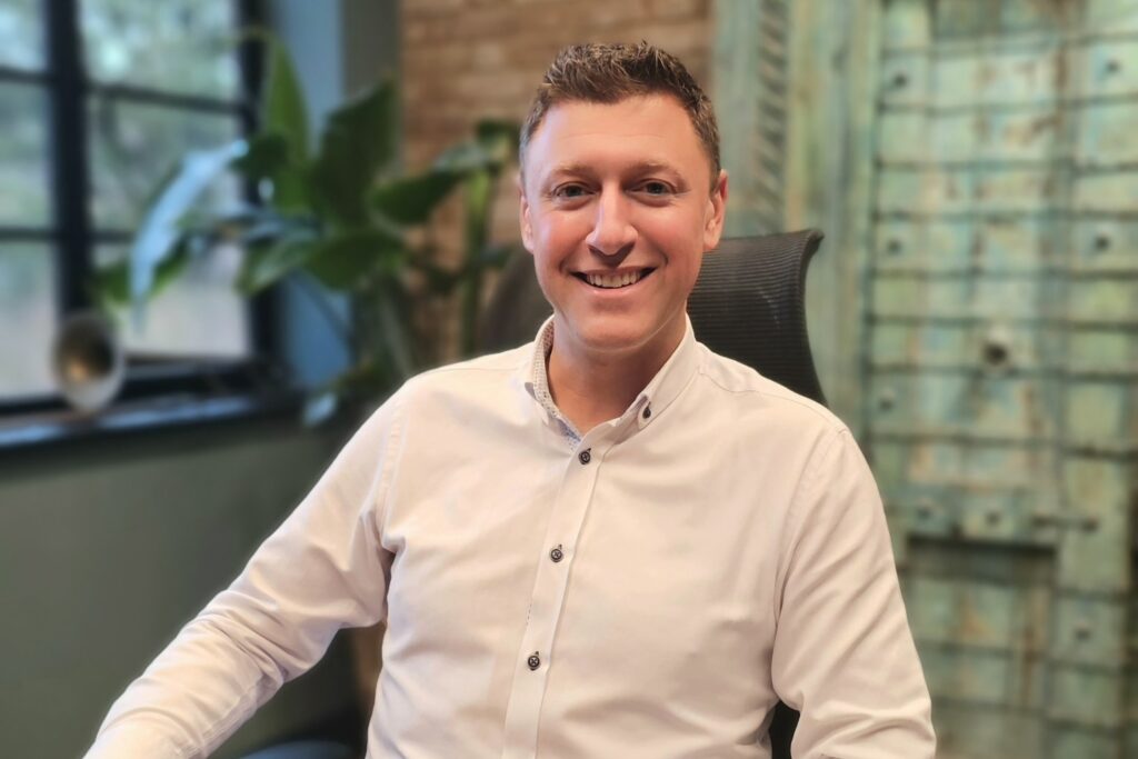 A man wearing a white shirt is sat at his desk , facing the camera and smiling