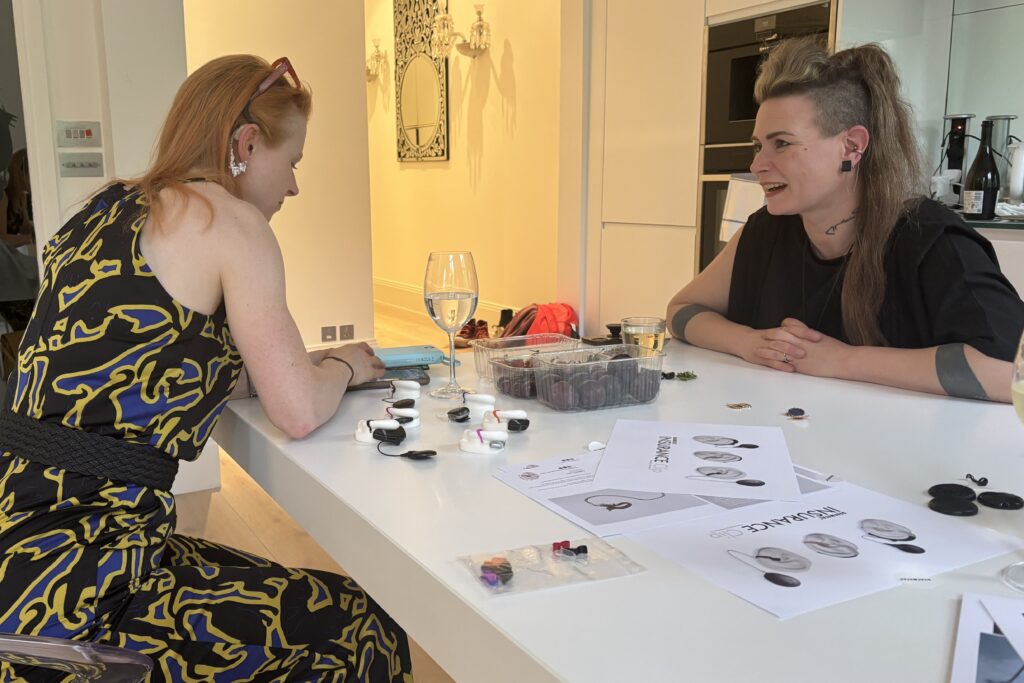 Two women sit across from each other at a white table, talking. Spread across the table are glasses, hearing instruments, hearing instrument jewellery and papers.