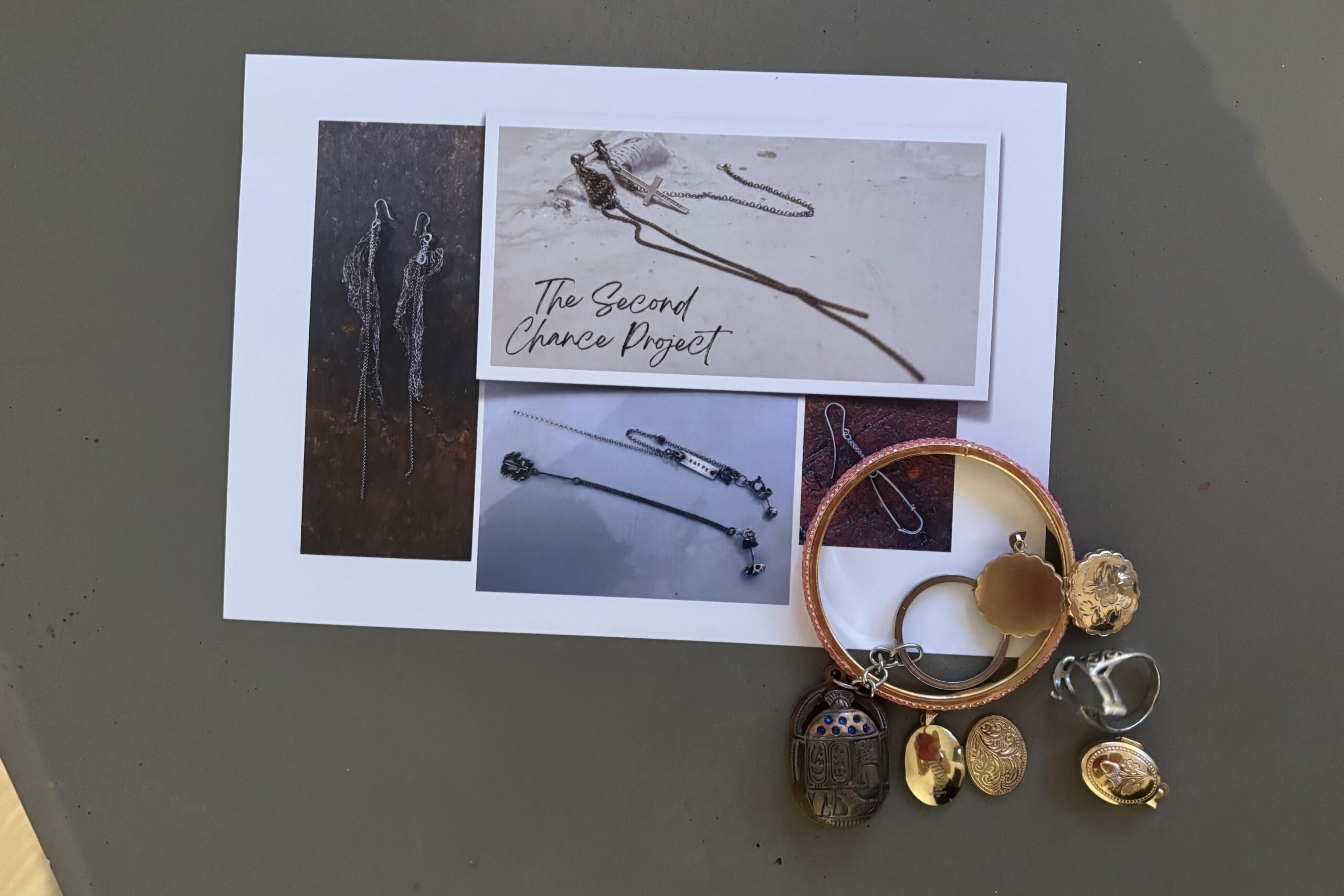 A close up photo of a small postcard with images of jewellery on it, along with the words 'Second Chance project' sits on top of a grey table, with a small pile of mixed jewellery pieces covering one corner of the postcard