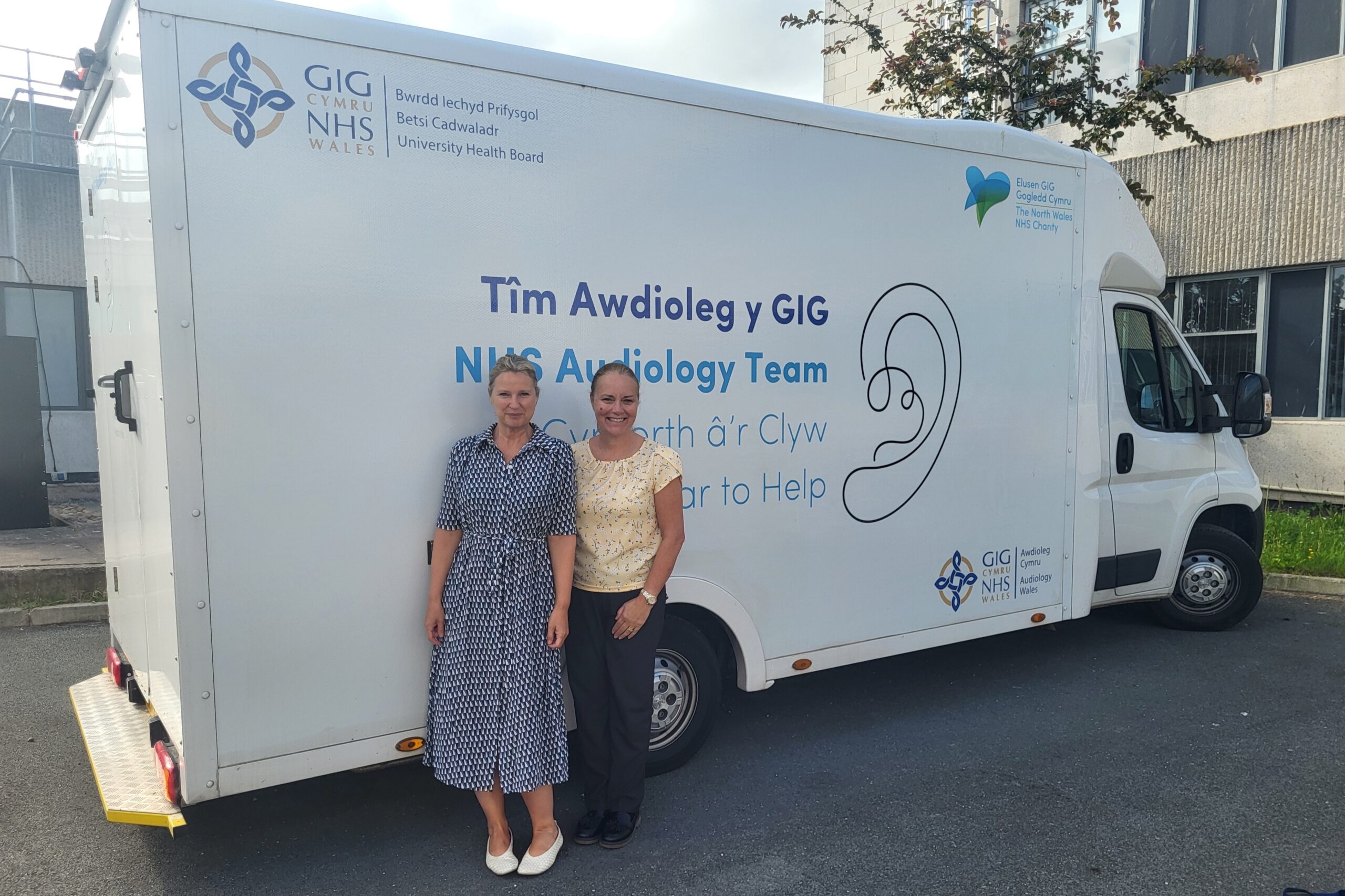 Two ladies stood smiling at the camera with their backs to a large white van