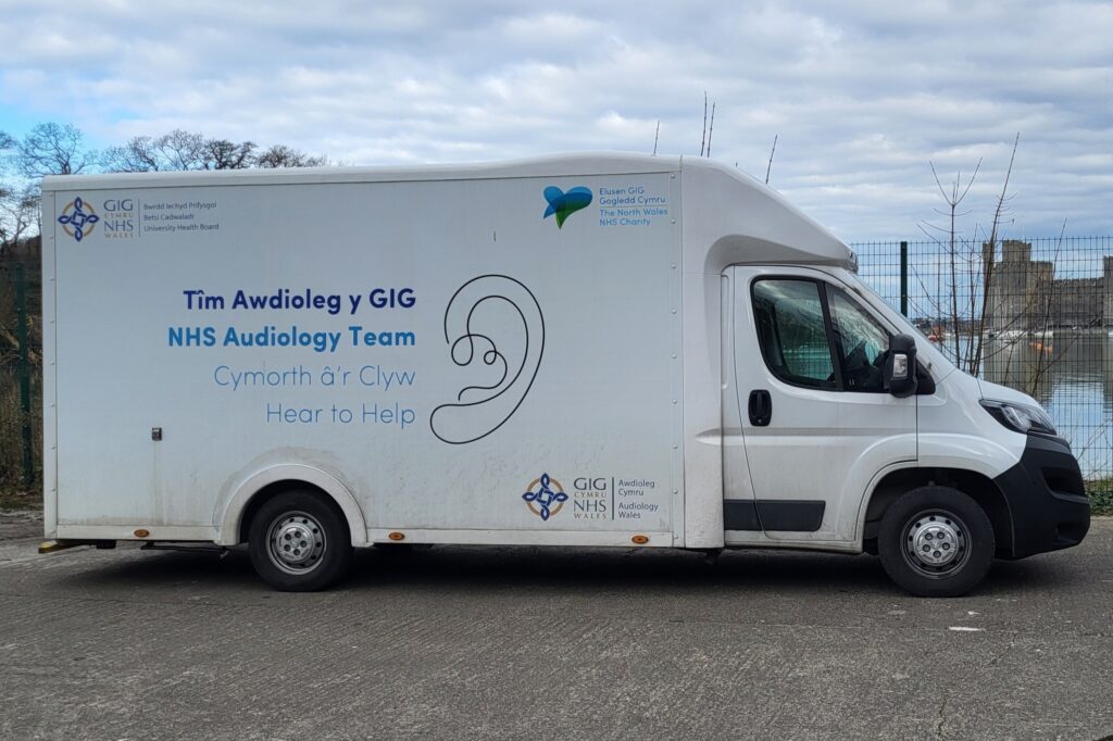 A side view of a large white audiology van, with text on the side of the van that says NHS Audiology team in English and Welsh