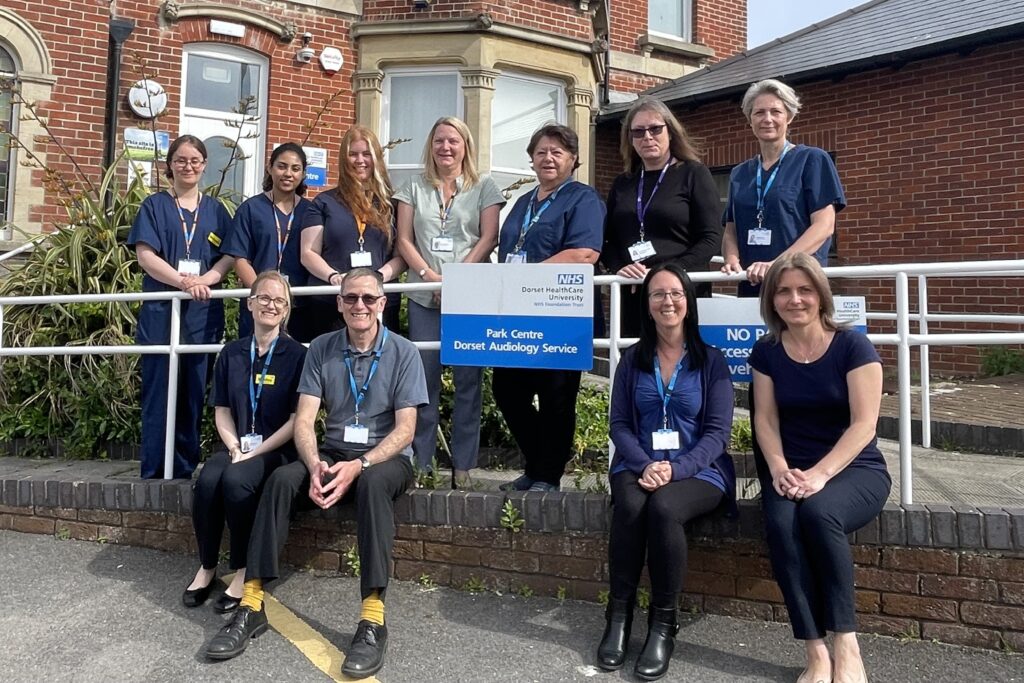 A group of 11 people, with four sat on a wall next to an NHS sign and seven other stood up behind the sign. all workers at an audiology unit